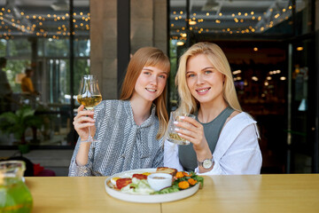 portrait of good-looking smiling caucasian women in restaurant, they sit together, hold glass of champagne in hands and look at camera