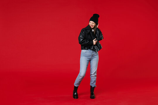 Full length of smiling cheerful beautiful young brunette woman 20s wearing basic black leather jacket white t-shirt hat put hand on head keeping eyes closed isolated on red background studio portrait.