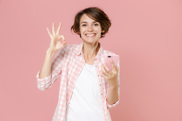 Smiling young brunette woman wearing casual basic checkered shirt standing using mobile cell phone typing sms message showing OK gesture isolated on pastel pink colour background, studio portrait.