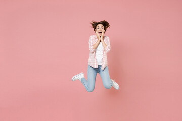 Full length of excited funny surprised young brunette woman 20s wearing casual checkered shirt jumping spreading legs put hands on cheeks isolated on pastel pink colour background, studio portrait.