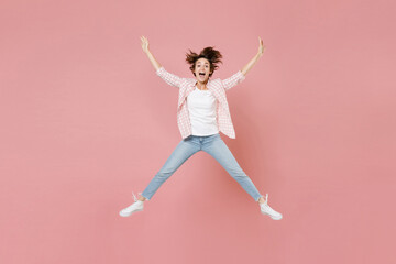 Full length of excited cheerful young brunette woman 20s wearing casual checkered shirt jumping spreading hands and legs keeping mouth open isolated on pastel pink colour background, studio portrait.