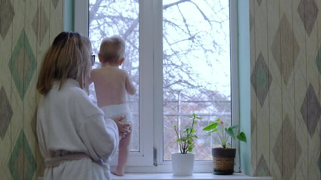 Disheveled Mom In A Dressing Gown Stands With A Child In Front Of The Window