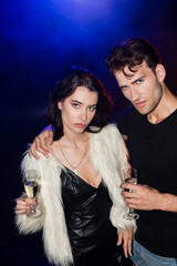 Serious man with glass of champagne embracing girlfriend in dress and white jacket with backlit on black