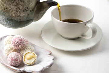 White coconut candy balls on handmade plate. Coconut cookies on white background with the cup of tea. Teapot with black tea. 