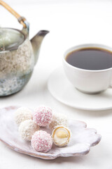 White coconut candy balls on handmade plate. Coconut cookies on white background with the cup of tea. Teapot with black tea. 