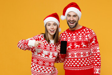 Excited young Santa couple friends man woman in sweater, Christmas hat pointing index finger on mobile phone with blank empty screen isolated on yellow background. Happy New Year celebration concept.