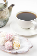 White coconut candy balls on handmade plate. Coconut cookies on white background with the cup of tea. Teapot with black tea. 