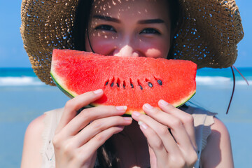 Happy beauty woman enjoy eating slice of watermelon on tropical beach in summer time. Attractive young asian woman standing on tropical beach in summer vacation time. Pretty female wear straw hat - Powered by Adobe