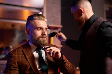 young hairstylist cutting hair to business man smoking in salon, relaxed male in elegant suit having hair care procedures