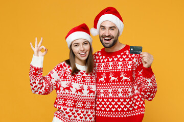 Funny young Santa couple friends man woman in red sweater Christmas hat hold credit bank card showing OK gesture isolated on yellow background studio. Happy New Year celebration merry holiday concept.