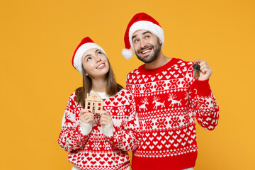 Pensive young Santa couple friends man woman 20s in red sweater Christmas hat hold house bunch of apartment keys isolated on yellow background studio. Happy New Year celebration merry holiday concept.