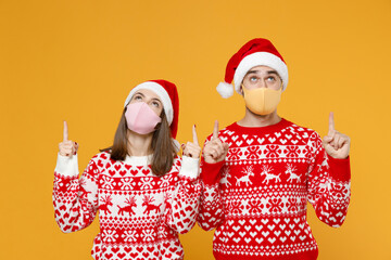 Young Santa couple friends man woman in sweater Christmas hat face mask safe from coronavirus covid-19 point index fingers up isolated on yellow background. Happy New Year celebration holiday concept.