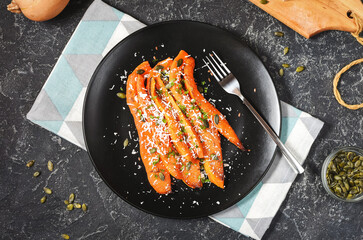 Baked slices of pumpkin with seasonings and feta on black stone background.
