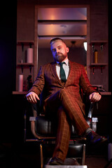 successful male barber shop owner sits feel himself superior, indoors, wearing formal suit or tuxedo, business people concept