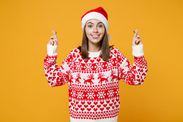 Excited young Santa woman in red sweater Christmas hat waiting for special moment, keeping fingers crossed making wish isolated on yellow background. Happy New Year celebration merry holiday concept.