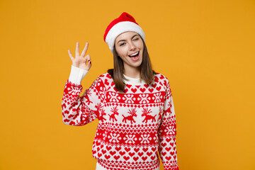 Blinking young brunette Santa woman 20s in red sweater Christmas hat showing OK gesture looking camera isolated on yellow background, studio portrait. Happy New Year celebration merry holiday concept.