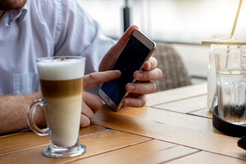 Fototapeta na wymiar A Caucasian man using smartphone while enjoying his morning caffe latte on top of wooden table. A man holding mobile with blank screen with coffee cup on wooden table. Man using phone in coffee shop.