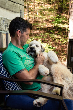 Man holding a fluffy dog on his lap outside on a deck in the woods.