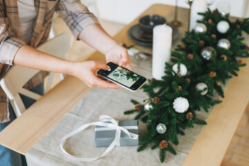 Young woman in plaid shirt takes photo gift box on festive table with the fir composition, christmas time