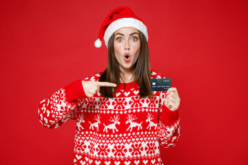 Shocked young Santa woman in sweater Christmas hat pointing index finger on credit bank card...