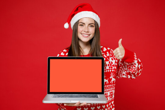 Smiling young Santa woman in sweater Christmas hat showing thumb up hold laptop pc computer with blank empty screen isolated on red background studio. Happy New Year celebration merry holiday concept. - Powered by Adobe