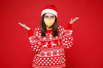 Irritated young Santa woman 20s in sweater Christmas hat face mask to safe from coronavirus virus covid-19 spreading hands isolated on red background. Happy New Year celebration merry holiday concept.