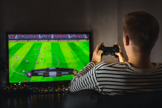 Young hipster man playing video game football soccer on console. gamer guy with gamepad controller holding Wireless joystick sitting on pouf at night. Back view. Fun entertainment at home isolation.