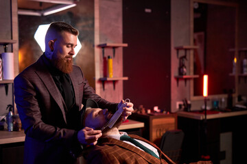 Fototapeta premium hairstyling process. barber cutting beard of caucasian male with the use of scissors, client sits in an armchair