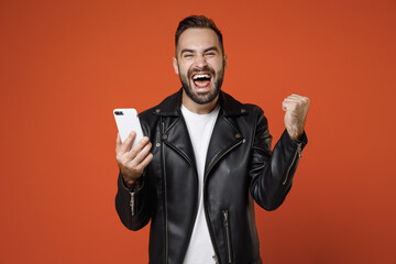 Happy young bearded man wearing white t-shirt black leather jacket standing using mobile cell phone...
