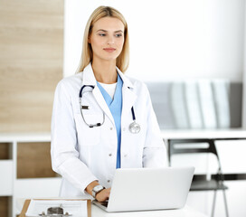 Blond female physician is lsmiling while using laptop computer. Woman-doctor at work in clinic excited and happy of her profession. Medicine concept
