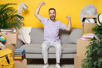 Happy owner man clenching fist like winner sits in living room on sofa at home household unpacking stuff in polythene indoor rents flat isolated on yellow wall. Relocation moving in apartment concept.