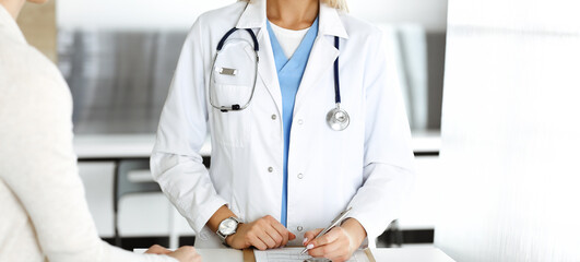 Unknown woman-doctor and patient in hospital. Blonde physician controls medication history records and exam results while using clipboard, close-up. Medicine concept