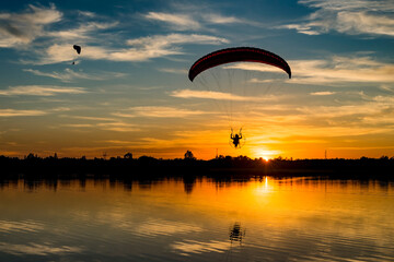 Two powered paragliders flying over the lake during sunset. Silhouetted paramotor pilot with...