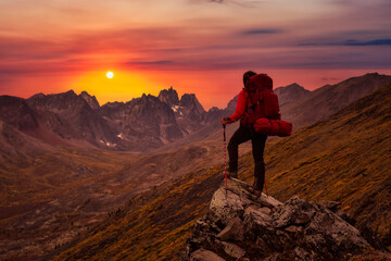 Fototapeta premium Woman Standing on Rocks looking at Scenic Mountain Peaks and Valley during Fall in Canadian Nature. Dramatic Twilight Sky Artistic Render. Landscape in Tombstone Territorial Park, Yukon, Canada.