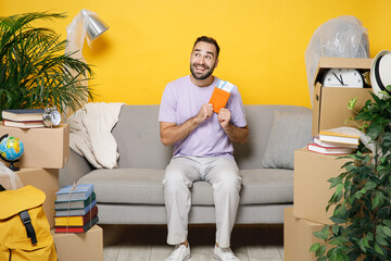 Pensive funny young owner man hold passport tickets looking aside sits in living room on sofa at home unpacking stuff indoor rents flat isolated on yellow wall. Relocation moving in apartment concept.