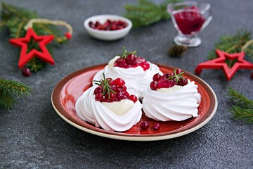 Portion Pavlova meringue pastries with cream and cranberry sauce in New Year's or Christmas decor on a red plate on a dark concrete background. New Year and Christmas desserts.