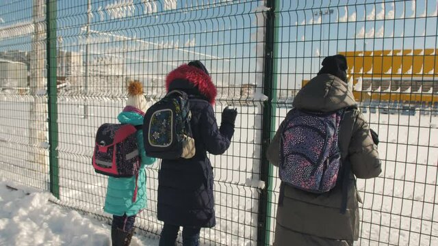 Three Schoolgirls in winter stand at the fence from the grid and look over the fence.