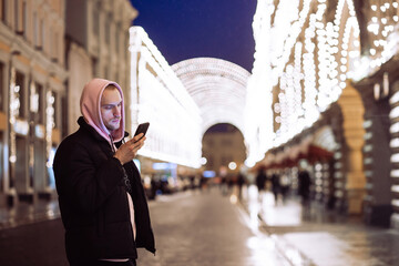 Man in hood looking into his smartphone in background of night street. Moscow. State Department Store