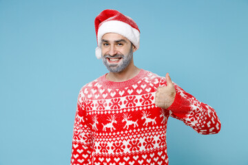 Funny Northern bearded man frozen snow face in Santa hat Christmas sweater showing thumb up isolated on blue background studio portrait. Happy New Year celebration merry holiday winter time concept.