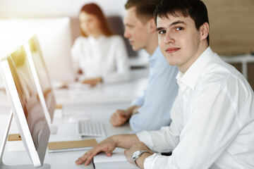 Fototapeta premium Cheerful smiling businessman at work in sunny office. Casual dressed entrepreneur using pc computer while sitting with diverse colleagues at the background