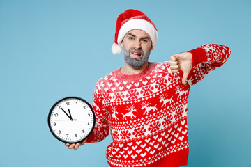 Displeased Northern bearded man frozen snow face in Santa hat Christmas sweater hold clock showing thumb down isolated on blue background. Happy New Year celebration merry holiday winter time concept.