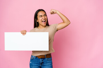 Young asian woman holding a blank paper for white something over isolated background raising fist...