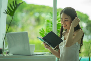 Beautiful young Asian woman in cozy pajamas reading a book while resting on the window sill at home.