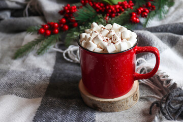 Obraz na płótnie Canvas Christmas hot drinks. Hot cocoa in a red mug with marshmallows on a wooden stand and background of gray cozy plaid. Winter cozy home concept. Close-up, selective focus, copy space