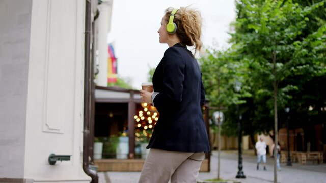 Modern business street office worker woman walking to a modern building holding digital tablet and coffee and wearing headphones