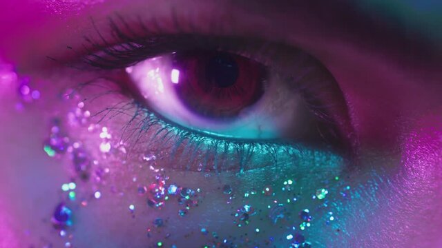 Female brown eyes in neon light. Young sexy girl in a nightclub. Macro look of the human eye. Fashionable glitter makeup on the face. Shining sparkles. Pink-blue-green color and new light, euphoria