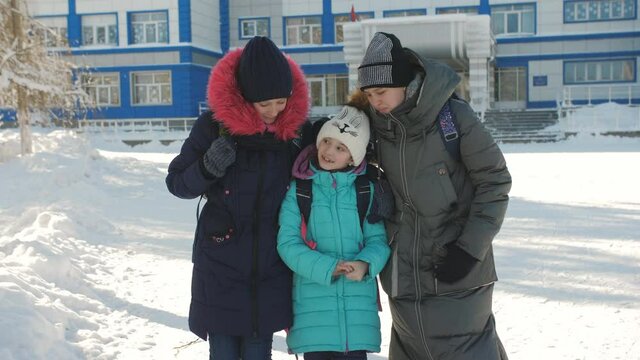 Two Schoolgirls Friends Pick Up Their Little Sister From School After School On A Sunny Winter Day.