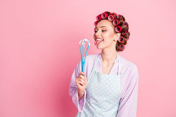 Photo of pretty house wife wear dotted blue apron red rollers looking holding licking whisk isolated pink color background