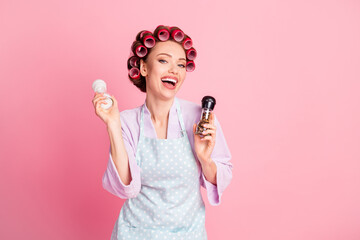 Photo of sweet house wife wear dotted blue apron red rollers holding two pepper-pots isolated pink color background