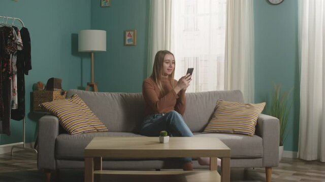 Young girl in blue jeans and a brown sweater sits on a beige sofa, looks at something on the phone and openly expresses joyful emotions